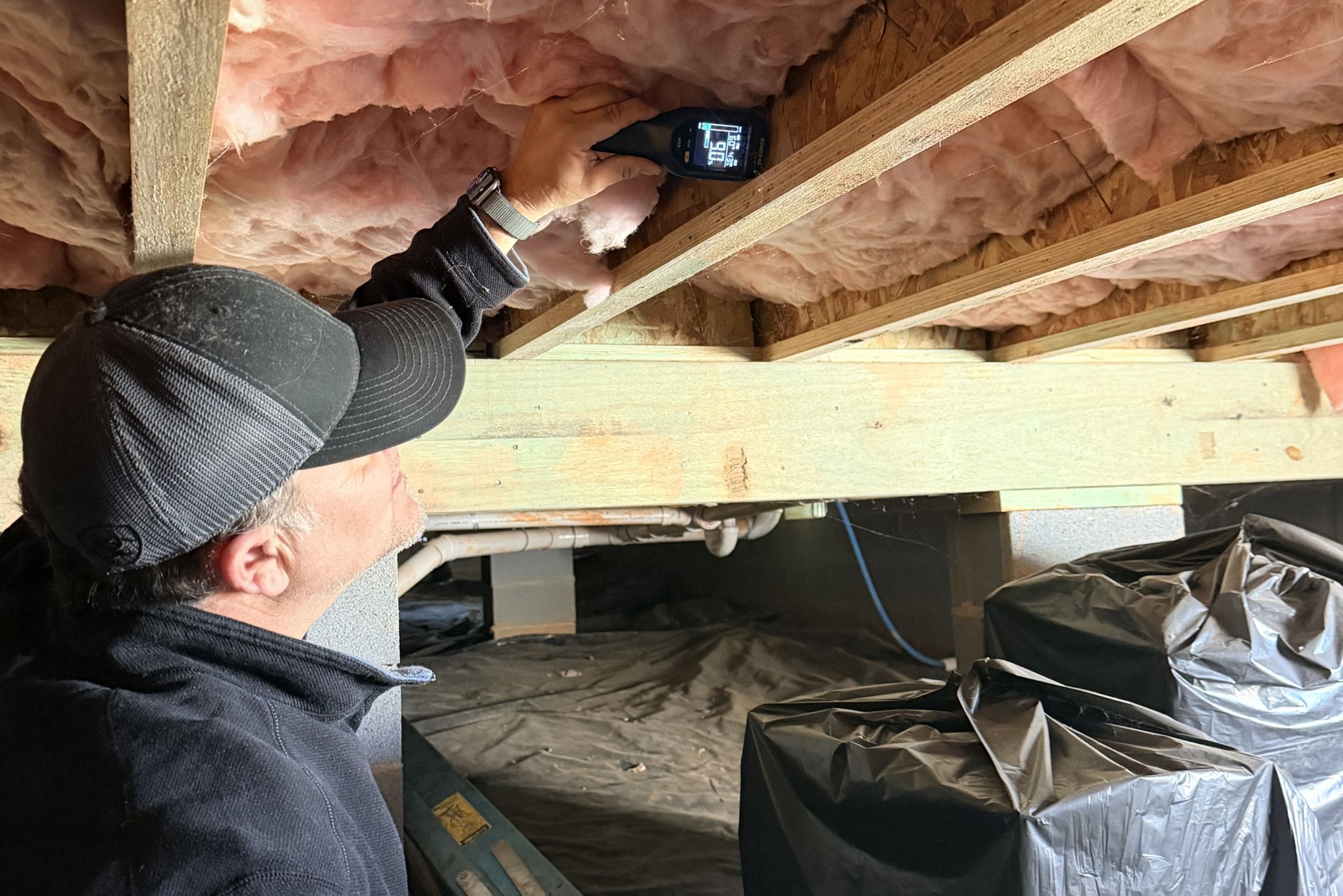 Technician in black cap using moisture meter on wood joist in crawlspace with pink insulation and black vapor barrier.