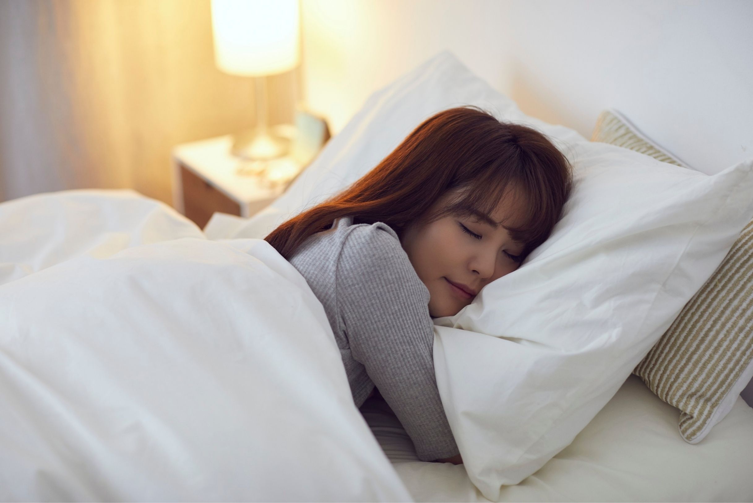 bed-bug-home-pest-defense-Fort-Mill-SC Woman with auburn hair in gray top sleeping peacefully under white duvet on white pillows with warm glowing bedside lamp.