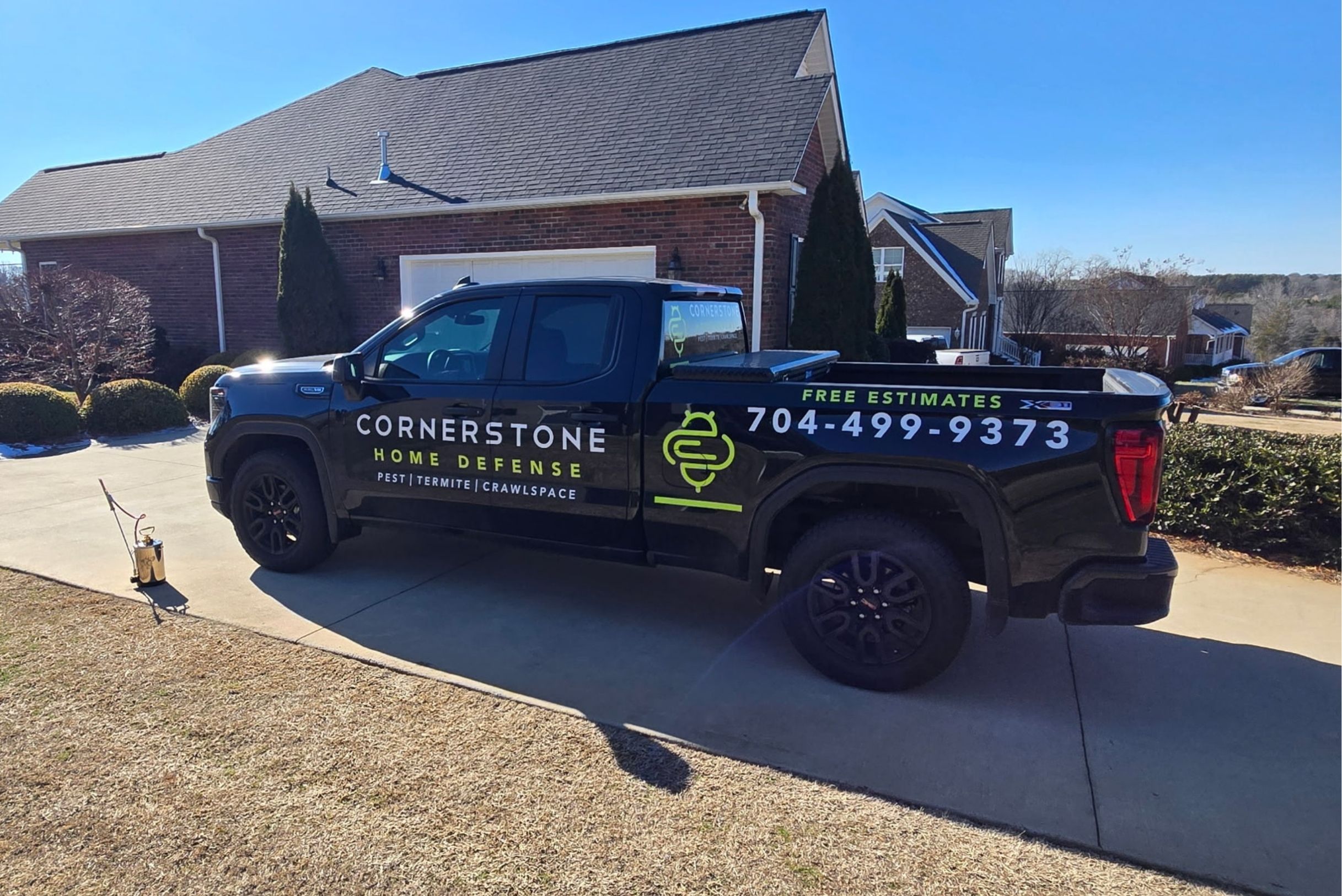 Black Cornerstone Home Defense GMC truck parked in driveway of brick home with pest sprayer on ground under blue sky.