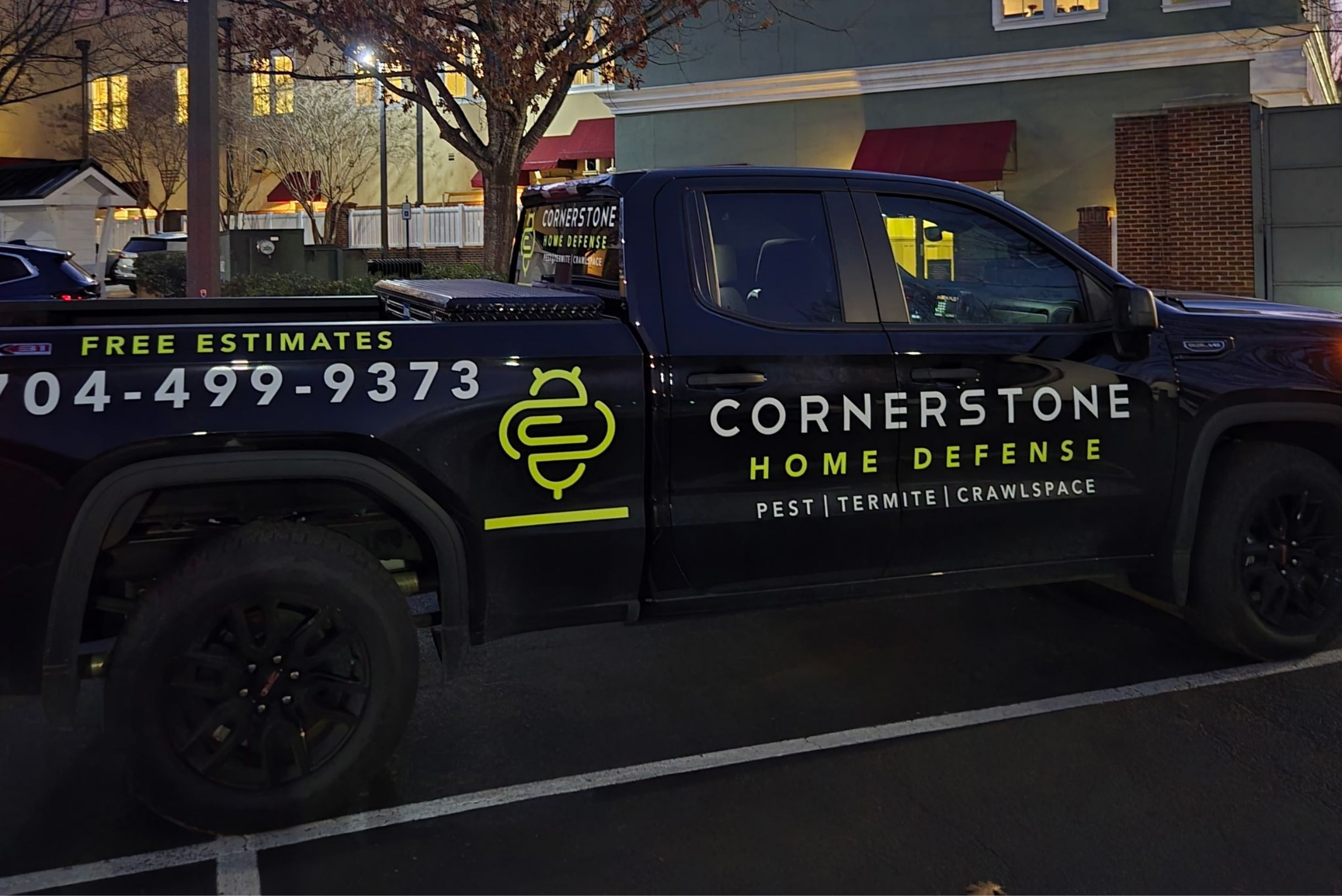 Black Cornerstone Home Defense truck with lime green branding and phone number 704-499-9373 parked in lot at night near brick building