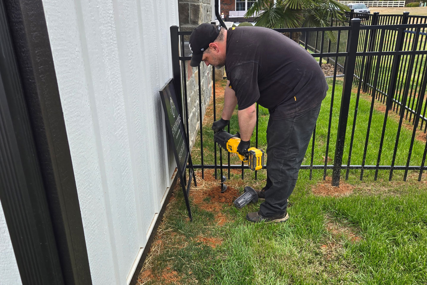 Technician in black cap drilling into red clay soil along white building foundation near black iron fence to install termite bait station.