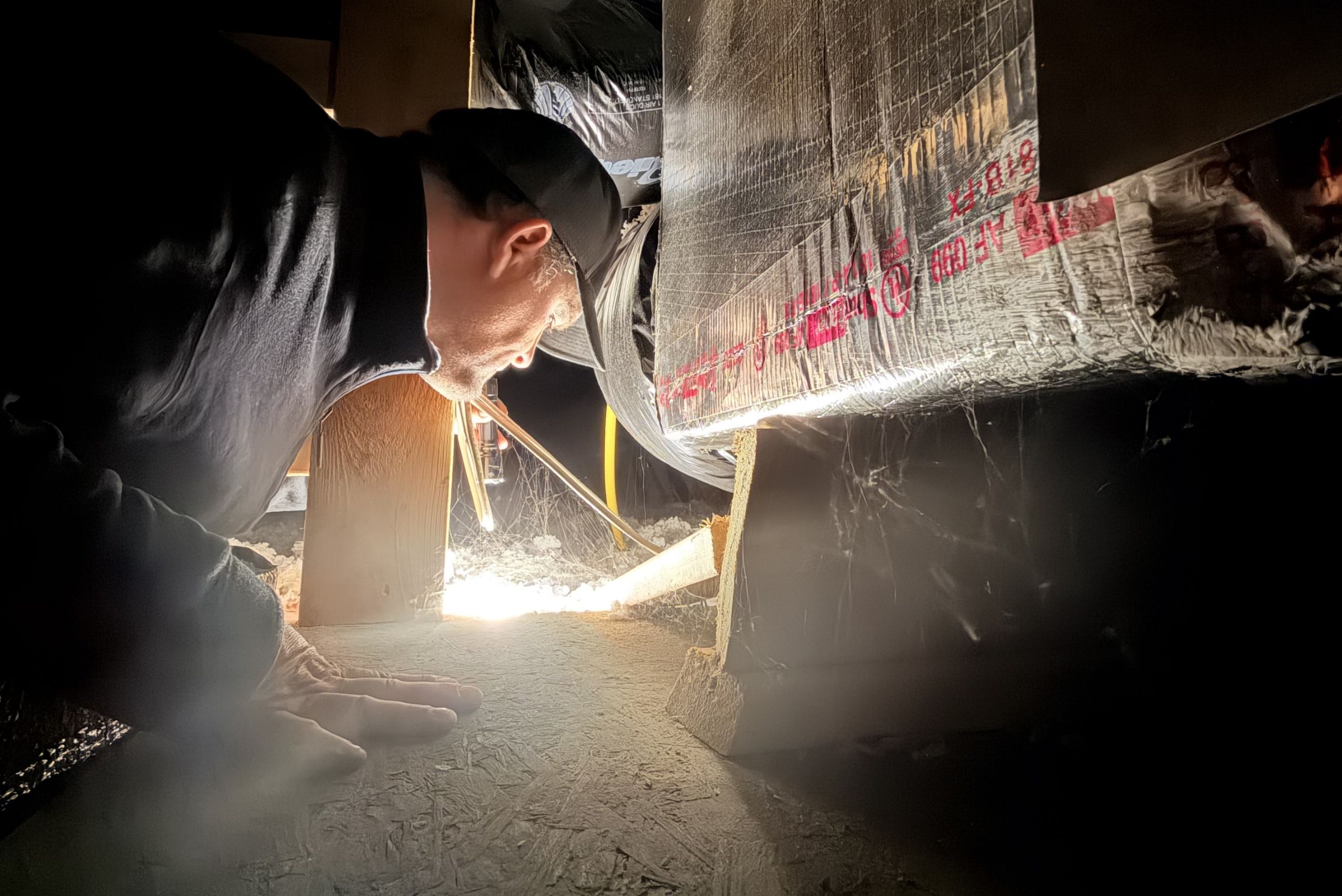 Technician in black cap crawling under dark crawlspace with bright work light inspecting silver insulation barrier with lots of webs.
