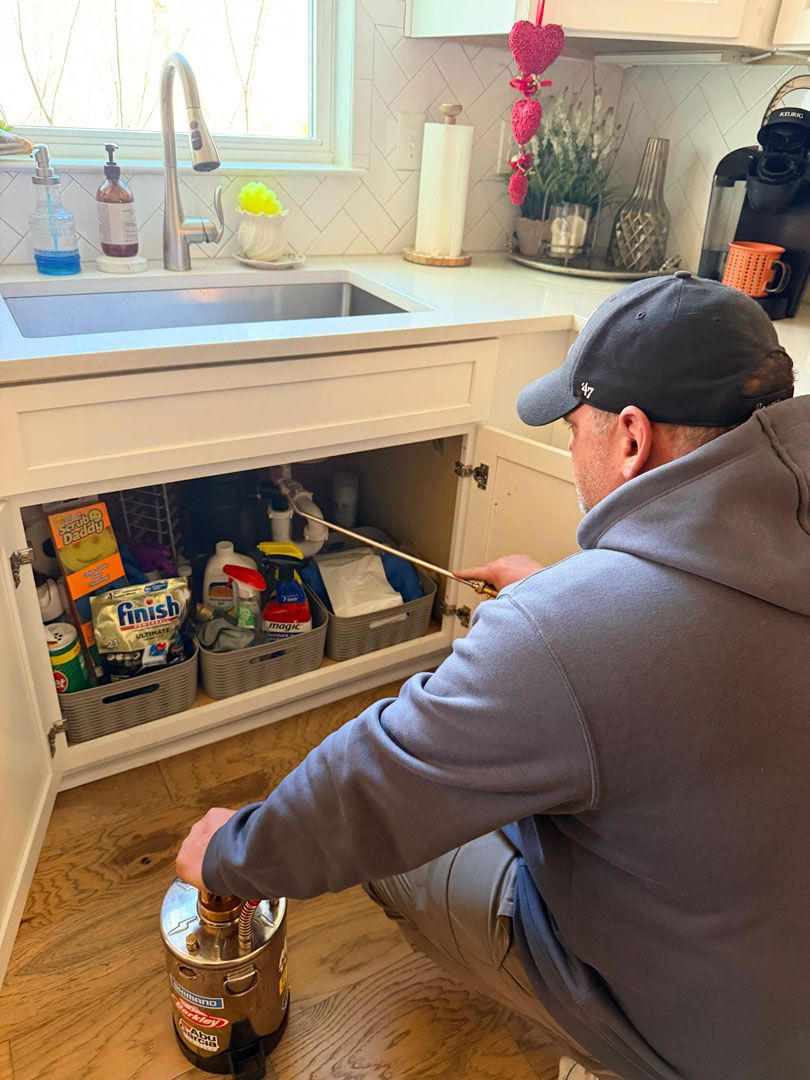 Technician in gray hoodie and black cap spraying copper wand inside open white kitchen cabinet under sink.