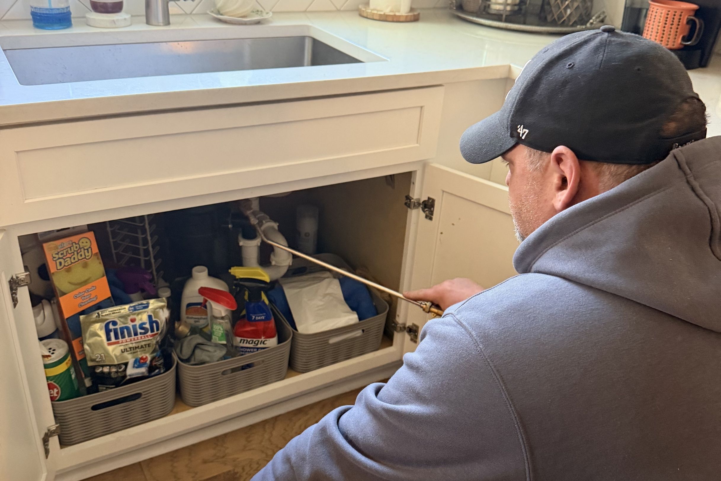 Technician in gray hoodie and black cap spraying copper wand inside open white kitchen cabinet under sink.