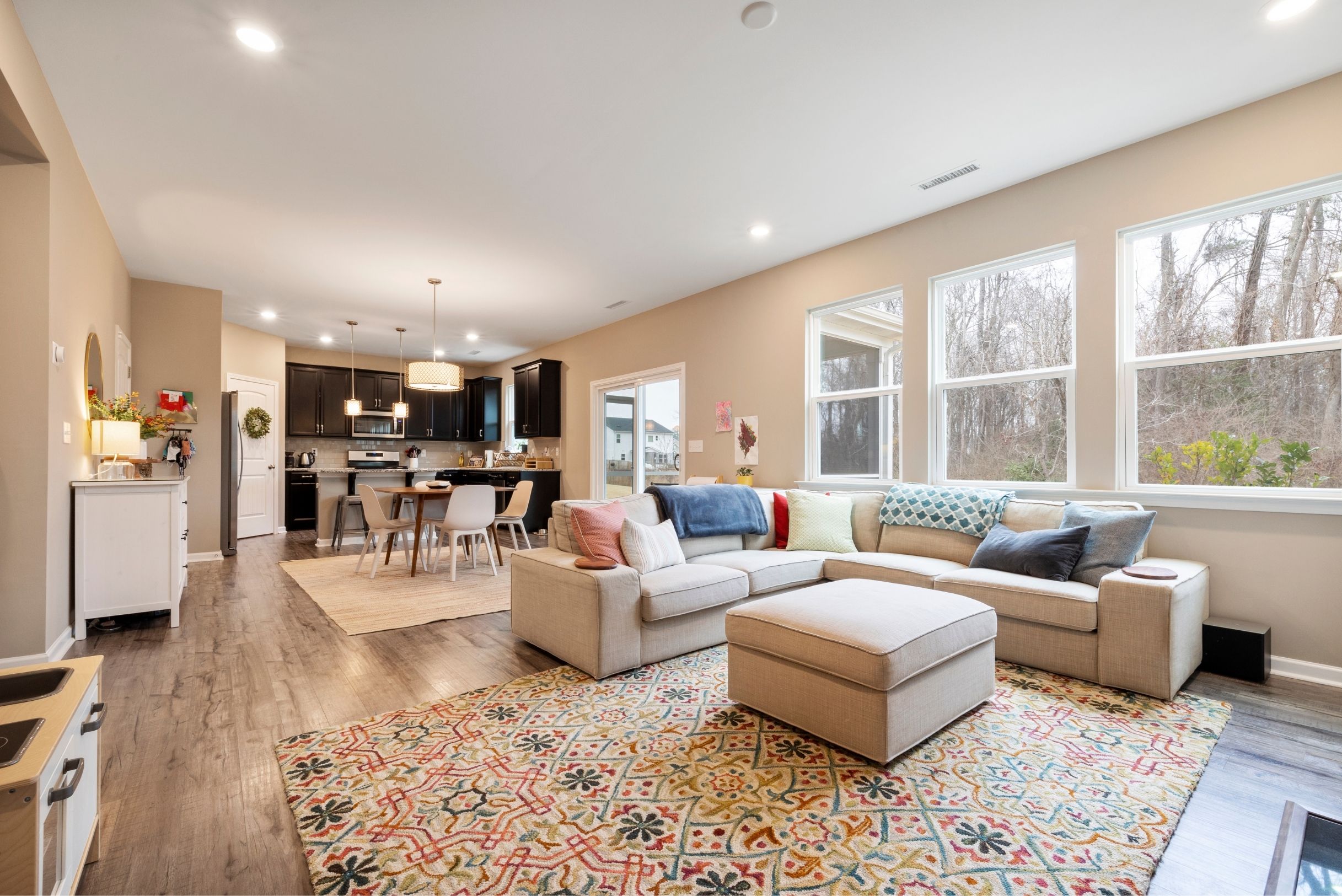 Open concept living room with beige sectional on colorful rug, dark kitchen cabinets and dining area in background.