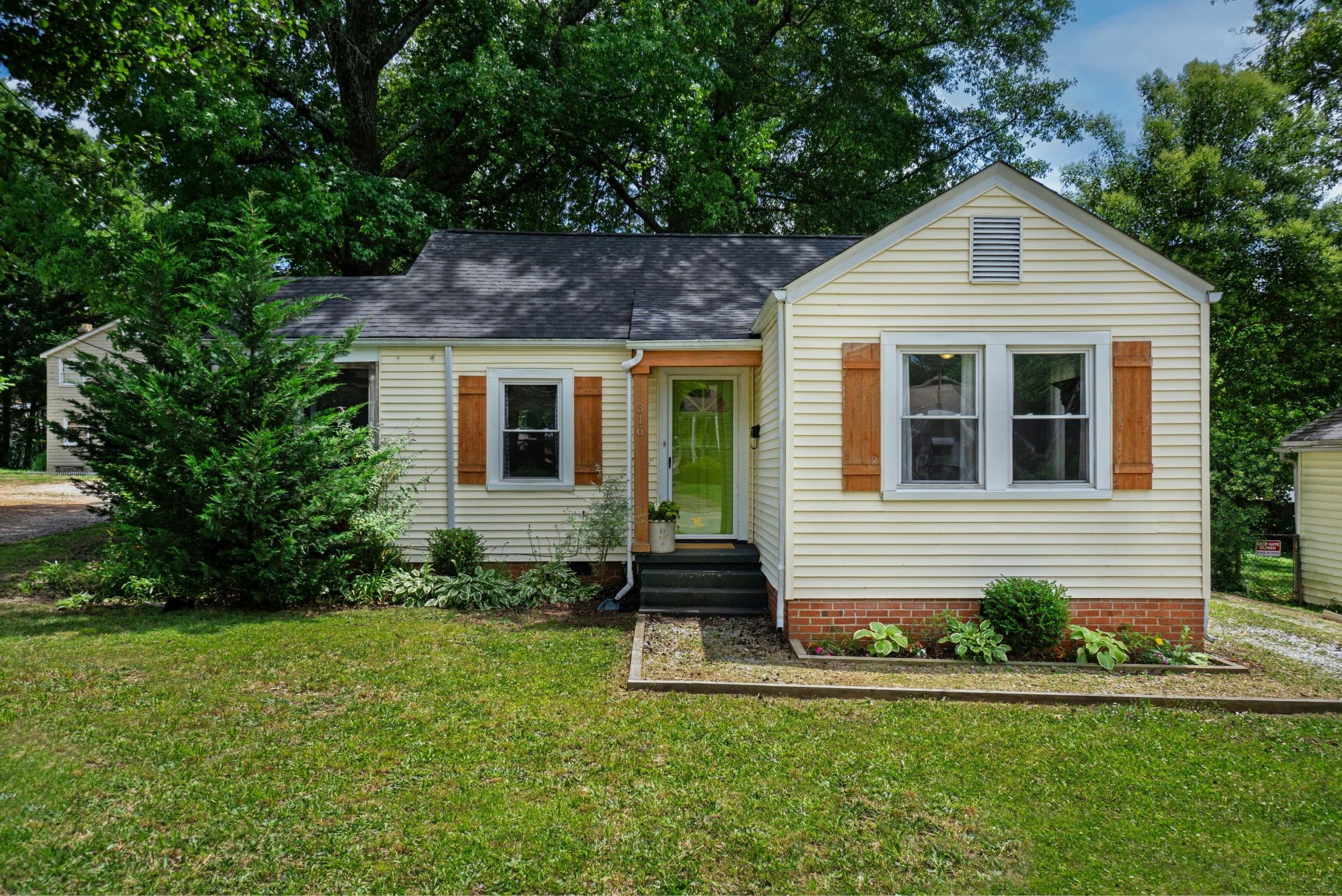 Cream ranch home with brown wood shutters, green front door, brick foundation, flower beds and large green trees.