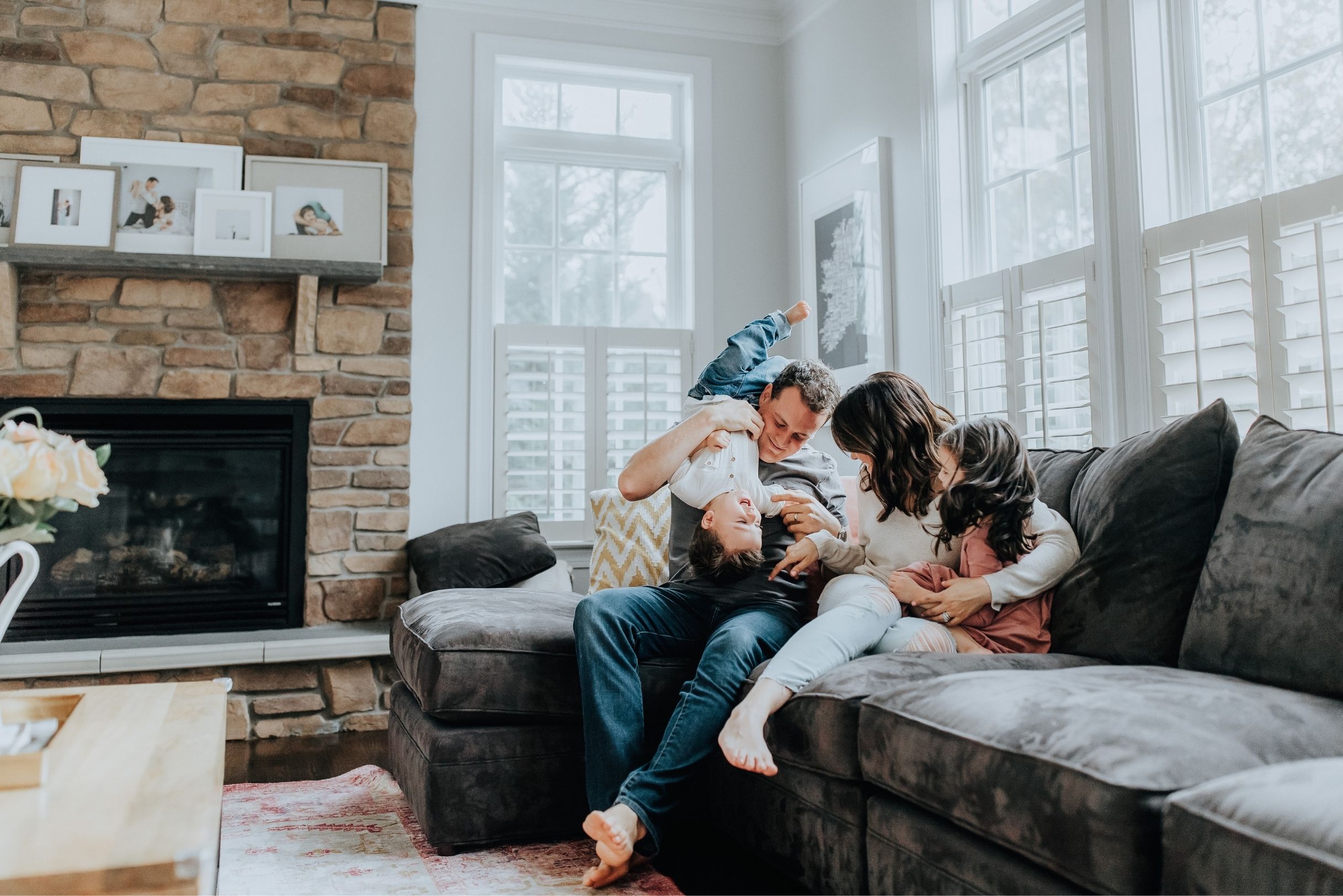 termite-control-in-fort-mill-sc Family of four laughing on gray sectional sofa in bright living room with stone fireplace and large white windows.