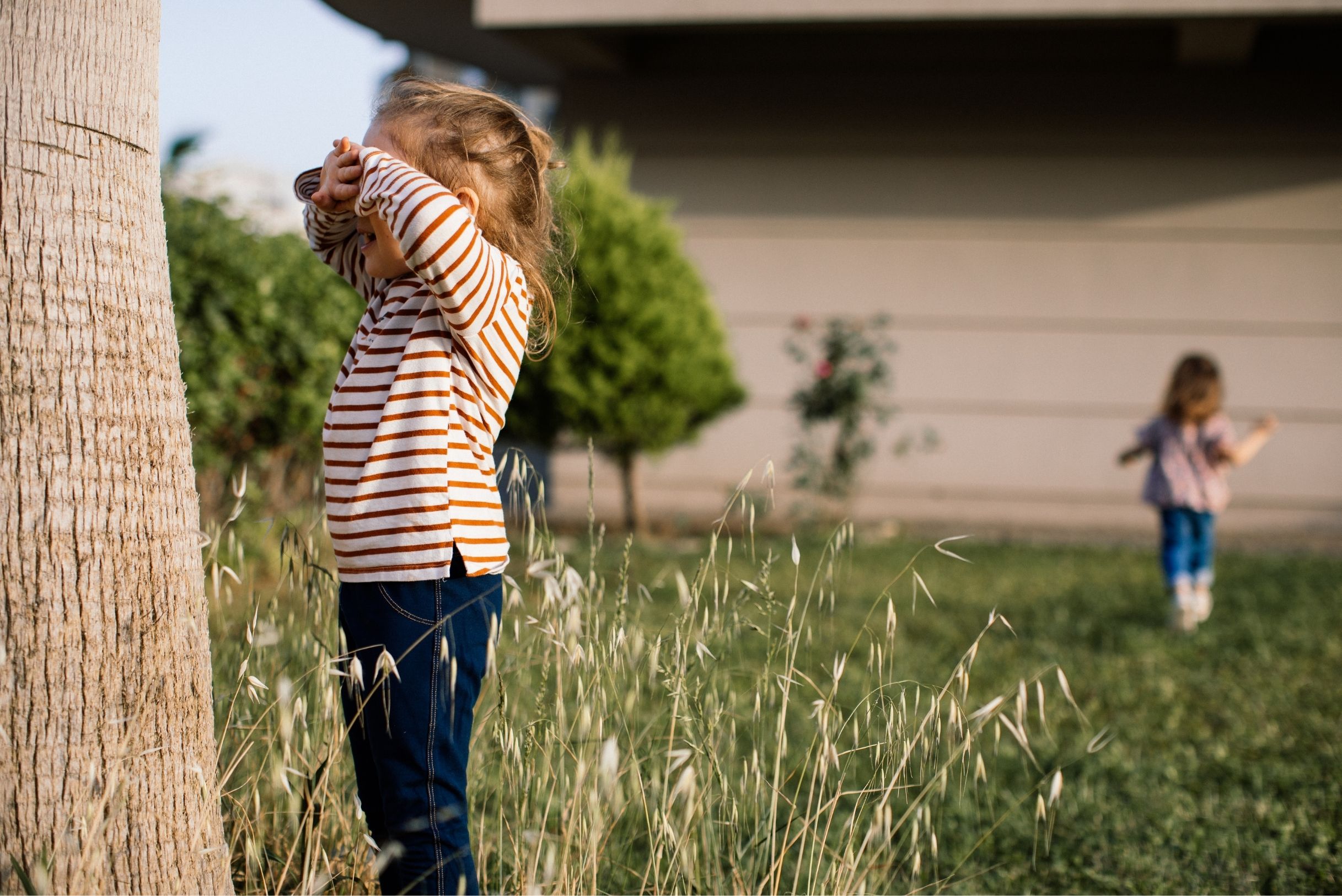 Girl in orange striped shirt covering eyes against tree trunk playing hide and seek with child in pink in grassy yard.