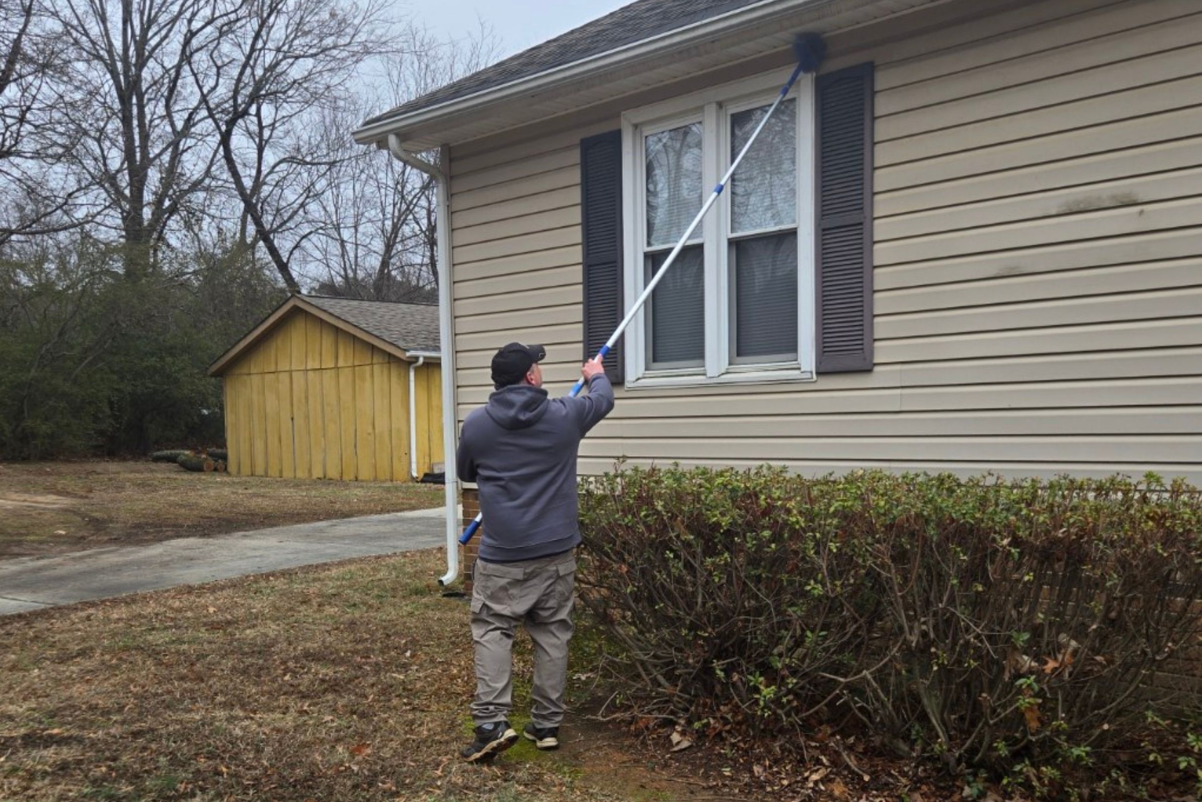 wasp-control-in-charlotte-nc Technician in gray hoodie using long blue extension duster to sweep cobwebs from roofline of tan house with dark shutters.