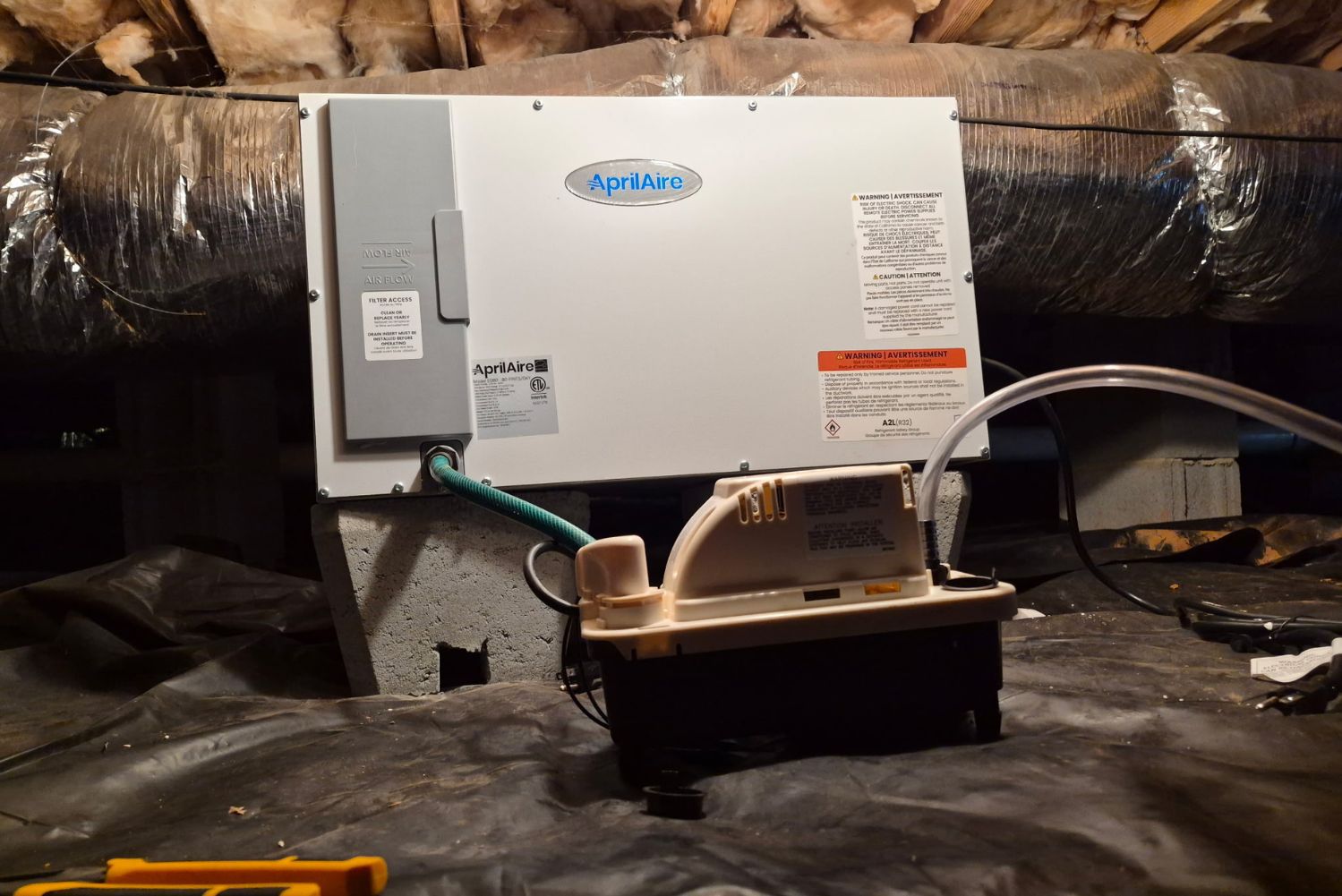 AprilAire dehumidifier unit mounted on concrete block wall with condensate pump and black vapor barrier in dark crawlspace.