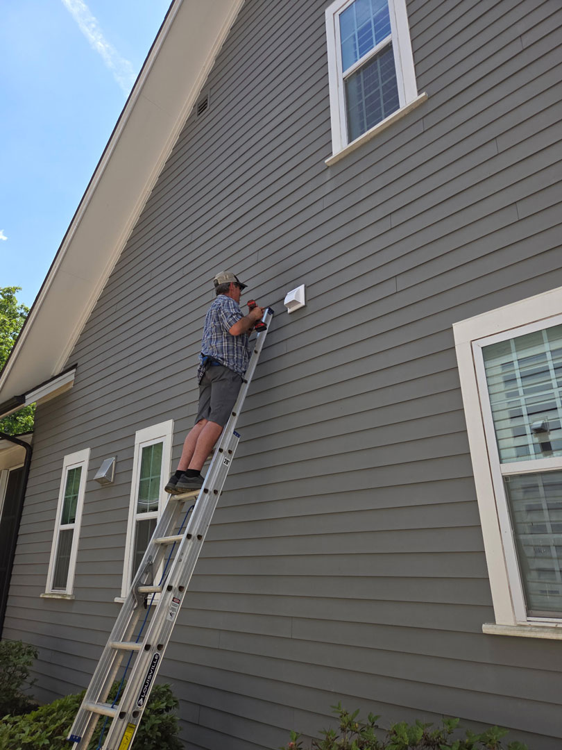 Cornerstone Home Defense technician on a ladder installing bird/bat exclusions on a grey home.